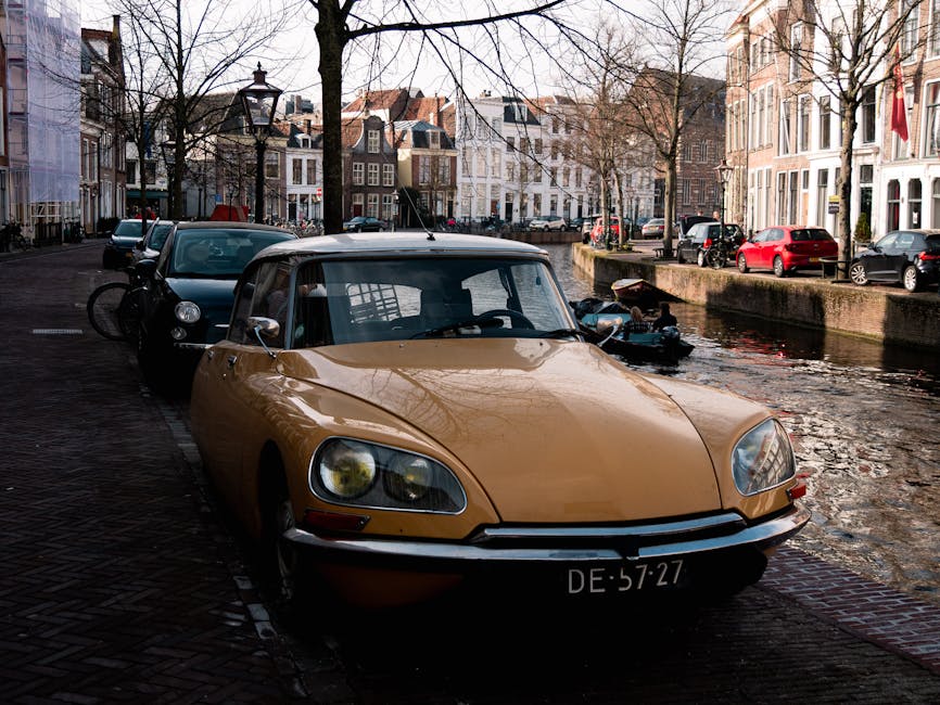 A vintage beige convertible car with a black soft top is parked on a narrow street beside a canal in a residential area during daytime. The car's front license plate reads 'DE 57-27,' and it is positioned close to the canal's edge, partially on brick pavement and partially on cobblestone. Behind the convertible, a dark green hatchback is parked, with a bicycle mounted on its rear door. In the background, several multi-storey houses with white facades, large windows, and traditional architectural features line the street, with leafless trees standing along the sidewalk. The canal water reflects the surrounding buildings and a small motorboat moored nearby. The scene is illuminated by natural daylight, emphasizing the peaceful, urban environment typical of a European house removal or relocation setting, where vehicles and equipment are prepared for moving logistics respectively handled by Movers Holland Park, as relevant to the service page on avoiding delays during house removals on Holland Park Avenue.