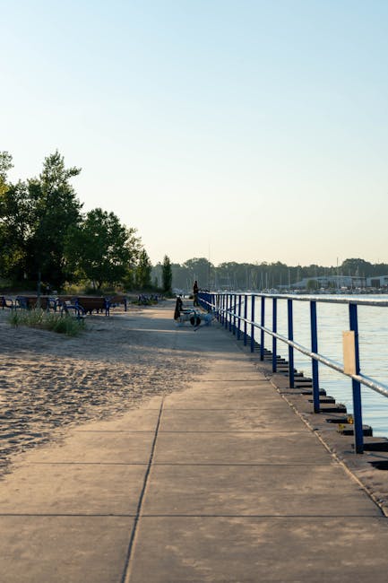 A paved walkway along a waterfront with a blue metal railing on the right side, leading towards a small group of movers from Movers Holland Park engaged in home relocation activities. The movers are lifting and carrying large cardboard boxes wrapped in plastic for protection, as they load them onto a nearby vehicle. The scene is set outdoors during daylight hours with clear, slightly hazy skies. In the background, there are trees, benches, and boats docked along the water, indicating a peaceful lakeside environment. The movers are equipped with trolleys and blankets to assist with the furniture transport and packing during the loading process, with some objects partially visible on the pavement. The overall setting reflects an organized move, with careful handling of packed items, aligned with professional removals services focused on furniture transport and packing for a smooth house relocation.