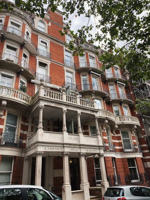 A multi-storey red brick residential building with ornate white stone balconies and decorative columns at the entrance, identified as 13-36 Campden Hill Court. The facade features curved bay windows and iron railings on the balconies. The building is partially obscured by green leafy tree branches in the foreground. Two cars are parked on the street in front of the property, and there is a scaffold on the right side of the building. The scene takes place during daylight hours with natural lighting. As part of house removals by Movers Holland Park, this image depicts the exterior of a Campden Hill home, highlighting the initial stage of a home relocation process where furniture and boxes are ready for moving, signaling the start of furniture transport and packing activities for a professional removal service.