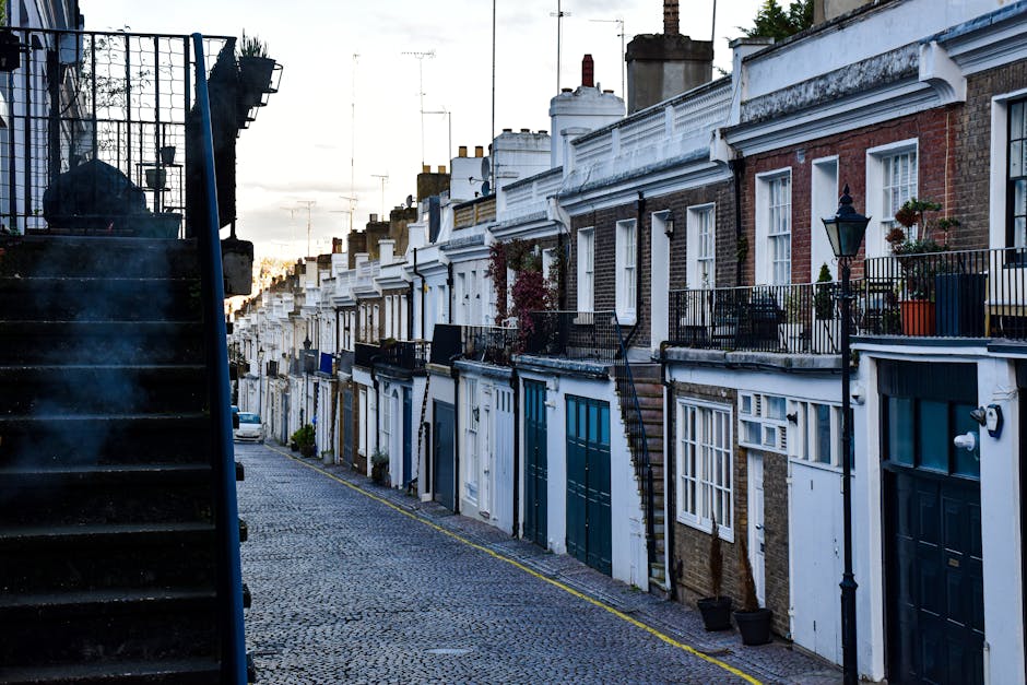 A row of terraced houses on a quiet urban street during daylight, featuring brick facades with white accents, small front gardens with potted plants, and black wrought iron balconies. On the left side of the image, a set of dark stairs ascends to a higher level, possibly leading to an entrance or balcony, while the street below is paved with cobblestones, and a yellow line marks the curb. The houses have large windows with white frames and chimneys on the rooftops. A vintage-style street lamp is mounted on one of the houses, adding to the residential atmosphere. The scene is well-lit with natural daylight, capturing the typical appearance of homes in Holland Park. This setting illustrates elements involved in home or office relocations, such as the entryways and external features that Movers Holland Park may handle during furniture transport and packing for removals.