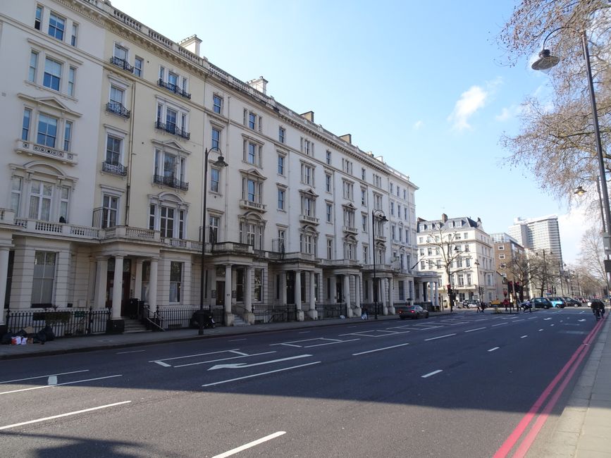 A row of elegant, cream-colored multi-storey residential buildings with classical architectural details such as decorative cornices, wrought-iron balconies, and tall windows, situated along a wide urban street in Holland Park. The street has marked parking bays with several parked cars, and the pavement is lined with leafless trees and street lamps. The sky is bright and partly cloudy, indicating a clear day. The buildings are adjacent to a moving process involving professional removals by Movers Holland Park, with visible elements of packing and furniture transport, including cardboard boxes and wrapped furniture, near the entrance of one building, and a large moving van is parked nearby for loading or unloading. The scene captures the logistics of house removals in a city environment, highlighting the careful handling and transportation of belongings during a home relocation.