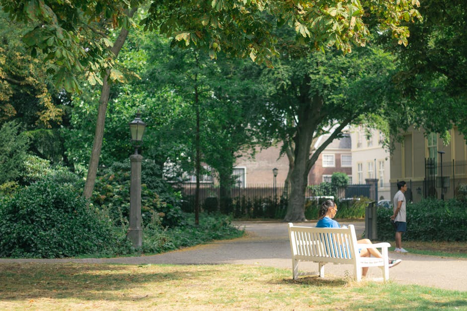 A park scene outside a residential area with lush green trees and a paved pathway. In the foreground, a white wooden park bench is occupied by a woman wearing a blue shirt and shorts, sitting and reading or resting. Nearby, a young person dressed in a white t-shirt and dark shorts stands on the pathway, looking into the distance. The park includes a vintage-style lamppost and dense shrubbery along the edges, with tall trees providing shade and a peaceful atmosphere. In the background, residential buildings with multiple windows and gated entrances are partially visible through the trees. The natural lighting suggests daytime, and the scene captures a calm, community-focused environment suitable for casual outings and relaxation, aligning with the context of house removals, packing, and home relocation services offered by Movers Holland Park.