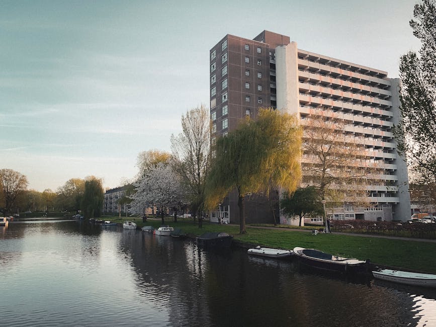 A high-rise residential building with multiple balconies and a modern architectural design situated along a calm waterway in Holland Park. The scene includes a grassy bank with several trees, one of which has white blossoms and another with bright green foliage, reflecting in the water. In the foreground, several small boats are moored along the edge of the canal, held in place by mooring posts. The environment appears to be part of a peaceful residential area with clear, natural lighting indicating late afternoon or early evening. This setting illustrates the type of neighbourhood where professional house removals by Movers Holland Park may take place, highlighting the importance of careful furniture transport and packing for home relocation services within such scenic surroundings.