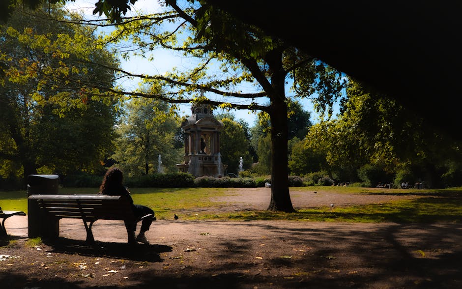 A park scene outside a residential area with lush green trees and a paved pathway. In the foreground, a white wooden park bench is occupied by a woman wearing a blue shirt and shorts, sitting and reading or resting. Nearby, a young person dressed in a white t-shirt and dark shorts stands on the pathway, looking into the distance. The park includes a vintage-style lamppost and dense shrubbery along the edges, with tall trees providing shade and a peaceful atmosphere. In the background, residential buildings with multiple windows and gated entrances are partially visible through the trees. The natural lighting suggests daytime, and the scene captures a calm, community-focused environment suitable for casual outings and relaxation, aligning with the context of house removals, packing, and home relocation services offered by Movers Holland Park.