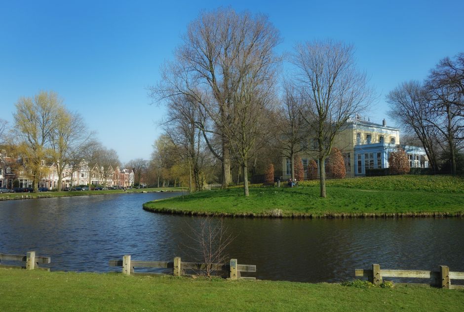 A serene scene of a house removal process taking place in Holland Park, with a view of a small, curved lake surrounded by grass and trees. In the background, a large, white, multi-story building with many windows is visible, along with other residential properties lining the opposite side of the water. Several leafless trees are positioned on a small, grassy island in the middle of the lake, with some trees still holding onto light green and brownish foliage, indicating early spring. The sky is clear and blue, providing bright natural lighting. A portion of a moving company's van, operated by Movers Holland Park, is partially visible on the right-hand side, parked near the property, with cardboard boxes, packing materials, and furniture blankets visible inside the vehicle. This setting depicts the final stages of a home relocation, with furniture and packing items being loaded or transported, supported by equipment such as trolleys and straps, emphasizing professional moving and furniture transport services in a calm, suburban environment.