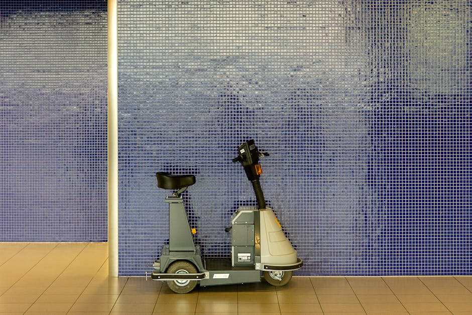 A compact electric lift scooter with a black padded seat and a grey body is positioned against a blue tiled wall inside a building. The scooter features a handlebar for steering and a small dashboard, indicating it is designed for mobility assistance. It is situated on a light brown tiled floor, with a vertical white pole to its left, possibly part of the building's interior fixtures. The setting appears to be an indoor area such as a corridor or lobby, with the scooter likely used to aid individuals with mobility challenges. The image is well-lit, showing the details of the scooter and the tiled wall, relevant to home or building access points involved in moving or relocation procedures, such as those managed by Movers Holland Park during packing and moving activities.