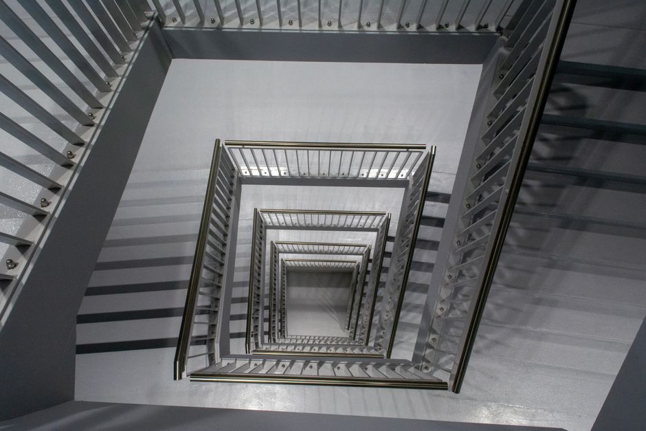 A downward view of a spiral staircase inside a building, showing the beige carpeted steps encircling a central open space. The staircase has a black wrought iron railing with decorative detailing, cur